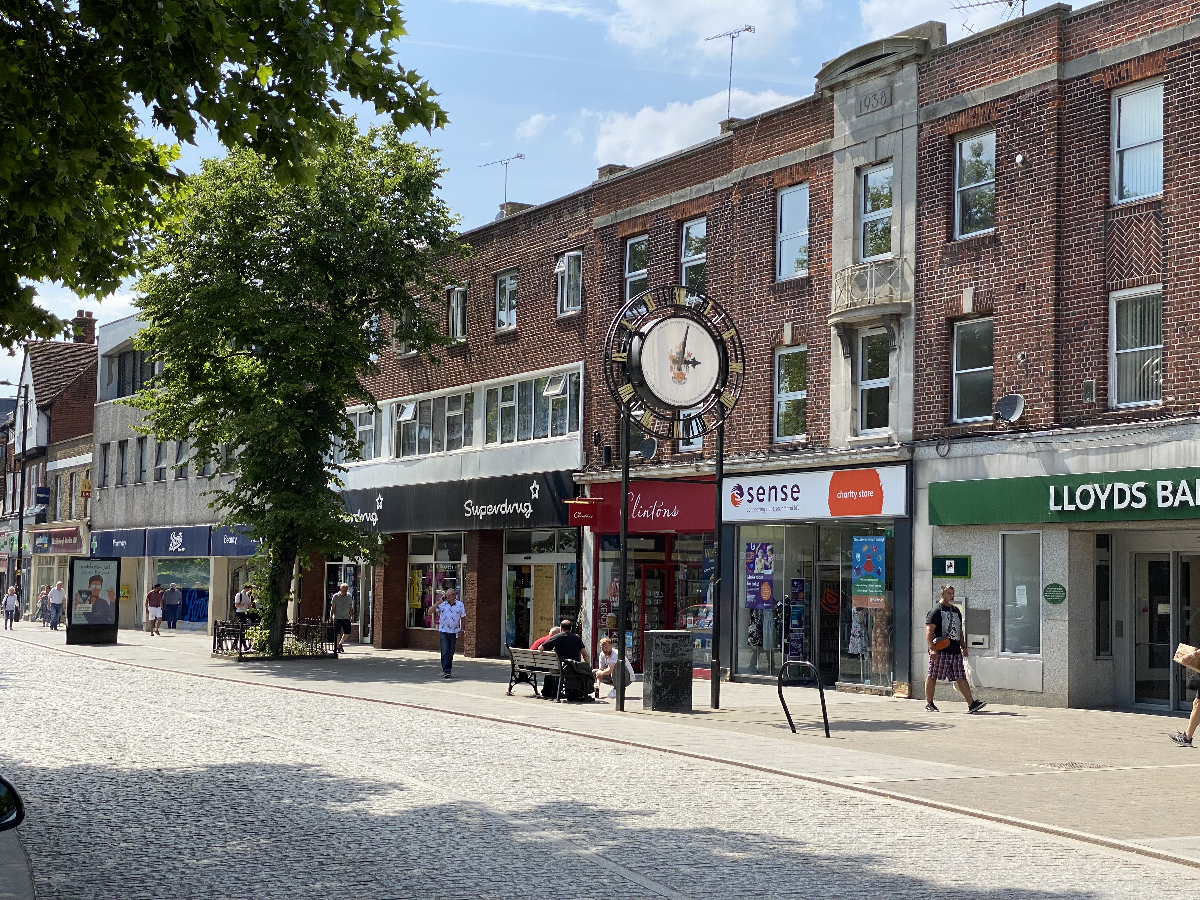 View of Brentwood High Street on a sunny day featuring the Brentwood Town Centre clock, trees and shops including Boots, Superdrug, Clintons, Sense charity store and Lloyds Bank