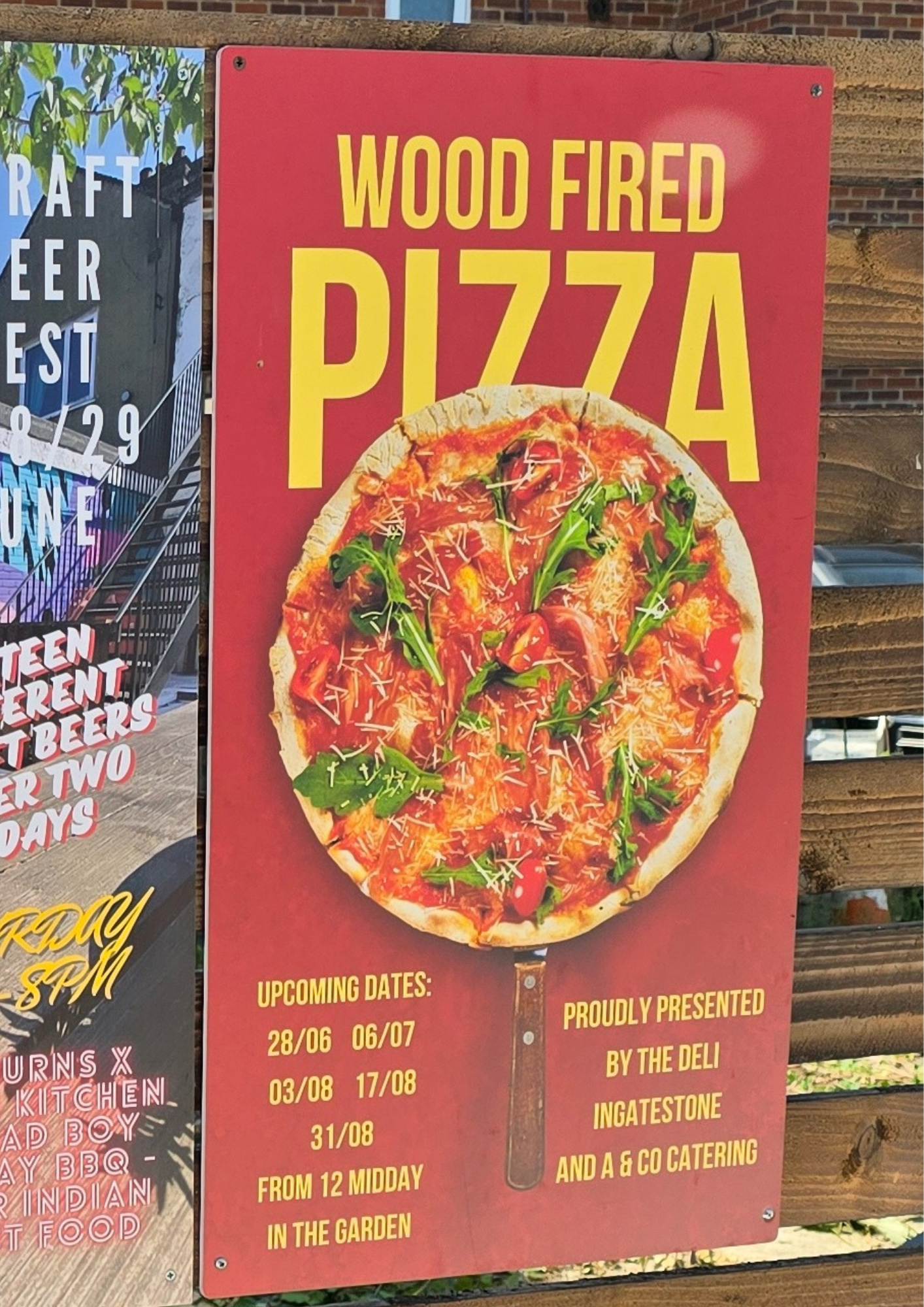 This image features a poster with the headline "Wood Fired Pizza" in large yellow letters on a red background. Below the headline is a photo of a wood-fired pizza topped with arugula, cherry tomatoes, and grated cheese. The poster lists upcoming dates for the event: June 28, July 6, August 3, August 17, and August 31, with each event starting at 12 midday in the garden. The event is proudly presented by The Deli Ingatestone and A & Co Catering.