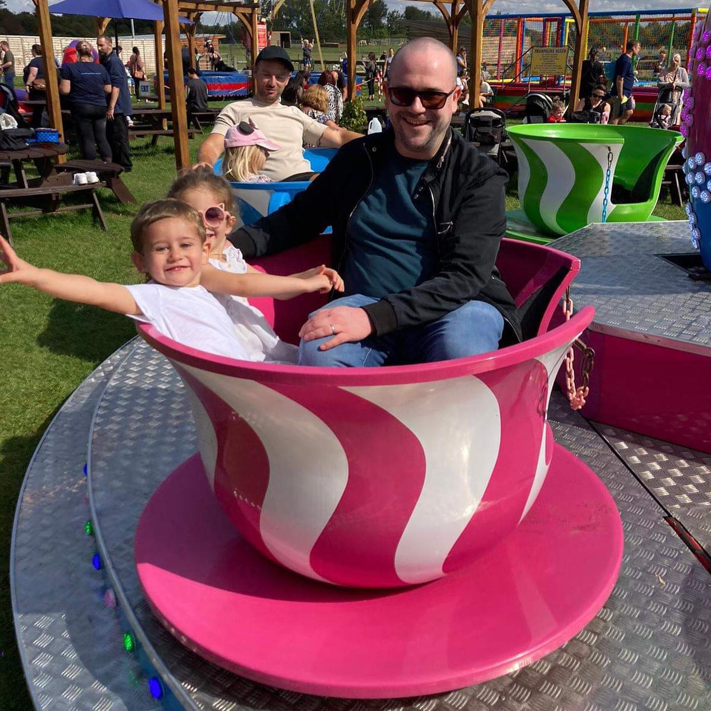A cheerful outdoor scene featuring a man and two young children enjoying a ride in a pink and white teacup at an amusement park. The children are smiling and appear excited, with one child stretching their arms out in joy. The ride is part of a teacup carousel, with other colorful teacups visible in the background. The area is lively, with other visitors sitting at picnic tables and enjoying the sunny day.