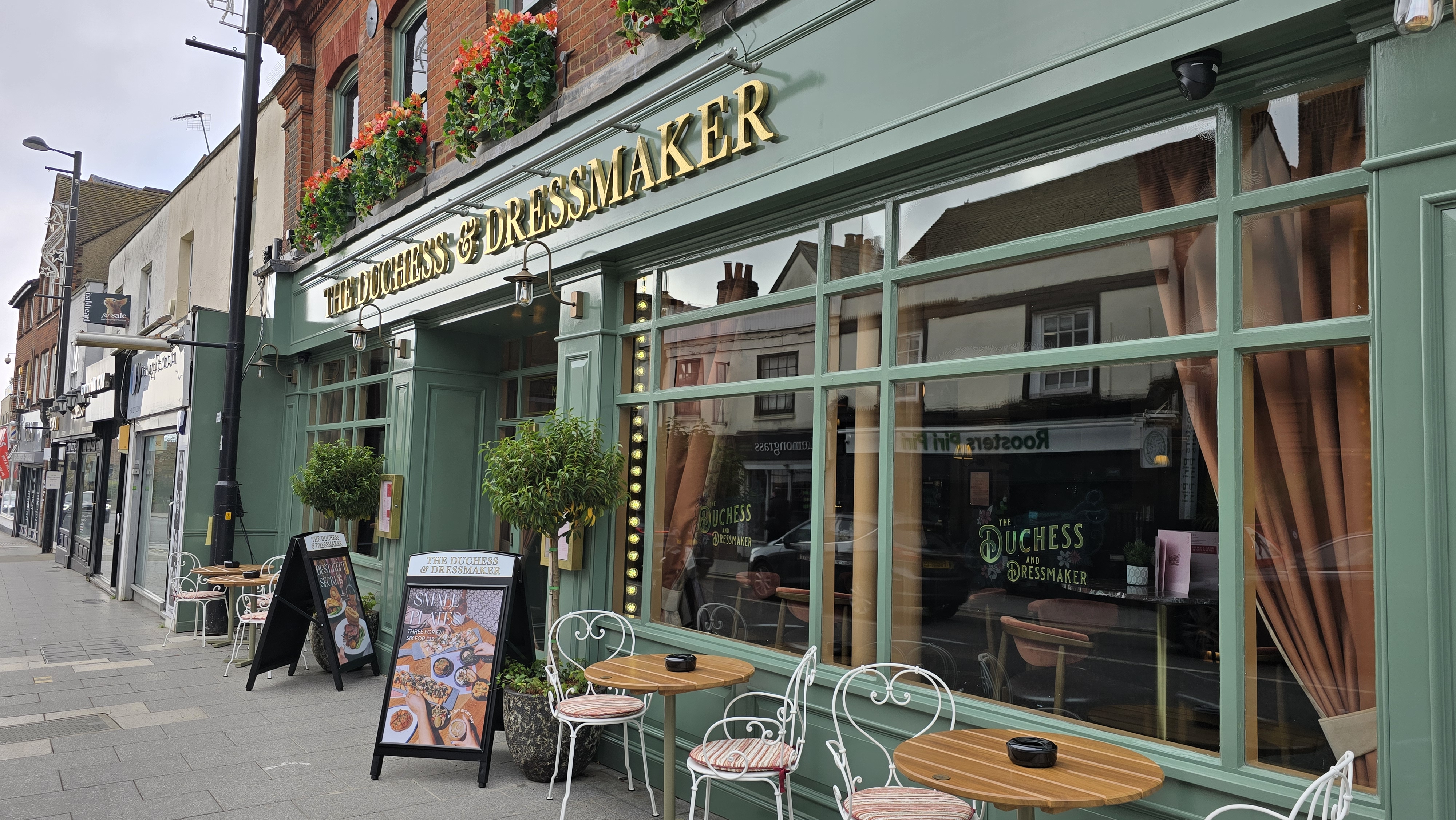 Image showing the exterior of The Duchess & Dressmaker, painted green with gold writing and velvet curtains at the windows. Two tables and four chairs are outside, while window boxes with red flowers are visible outside the first floor windows.