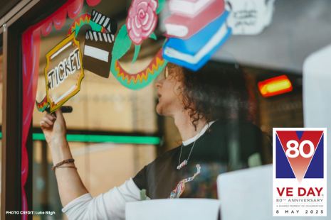 Image showing artist Sue Mills decorating a window with a colourful design.  The VE Day 80th Anniversary branding is in the bottom right corner.