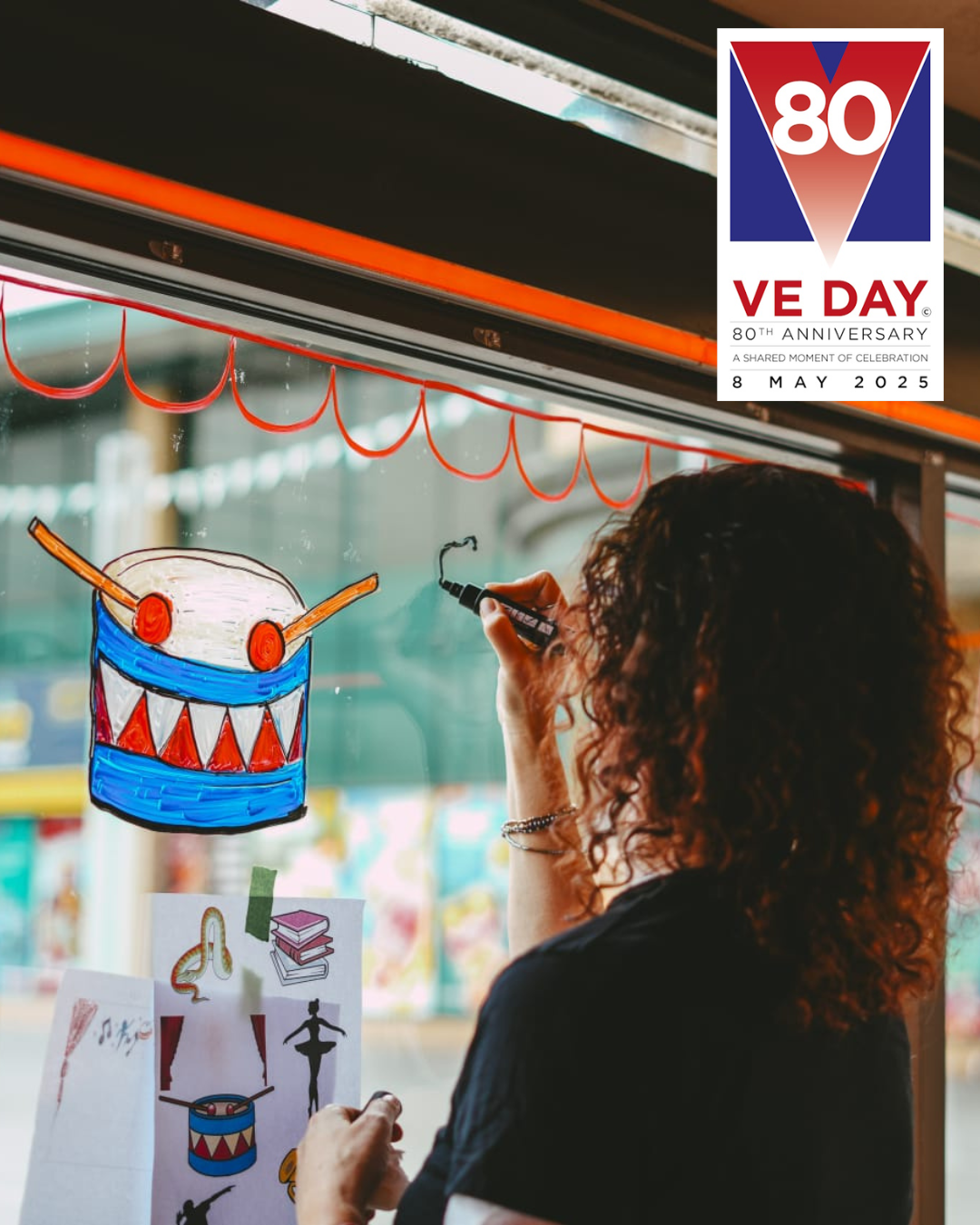 Image of artist Sue Mills decorating a window, with the VE Day logo in the top right corner.