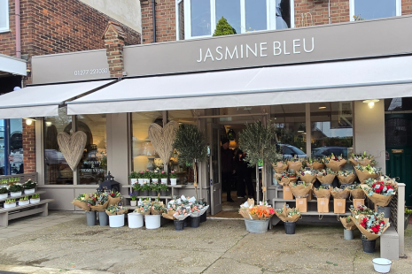 Exterior image of Jasmine Bleu florist in Shenfield - with a taupe sign and white text.  There are flowers arranged outside the shop with a large wicker heart on the left window.