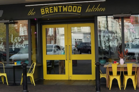 Exterior photograph of The Brentwood Kitchen with yellow text, yellow double doors and yellow chairs.